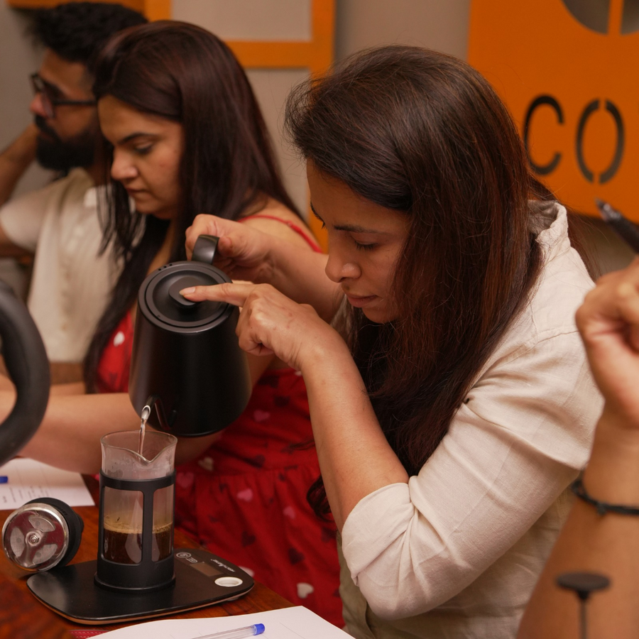 Two women preparing coffee at a table with a focus on one woman pouring water from a black kettle into a french press.