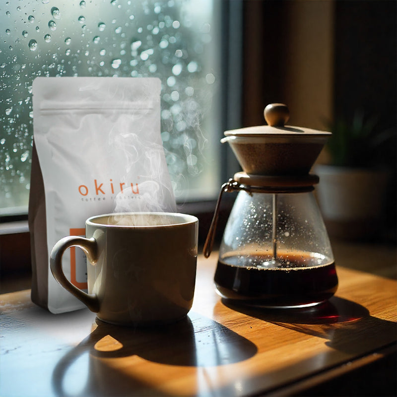 Cup of coffee, coffee maker, and okiru coffee bag on a wooden table with rain outside the window.