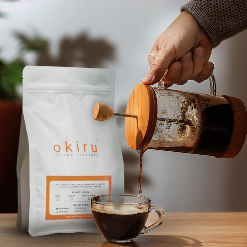 Person pouring coffee from a French press into a glass cup next to an okiru coffee bag.